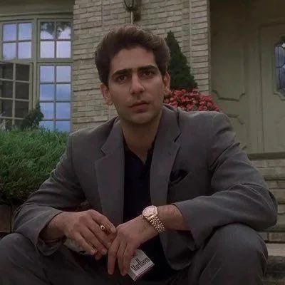A young man in a gray suit jacket sits on steps in front of a house with white siding, looking directly at the camera with a serious expression while holding a Marlboro cigarette pack and wearing a gold watch. The post humorously references the iconic "Sopranos" imagery of Christopher Moltisanti to playfully inquire about joining a club or group.