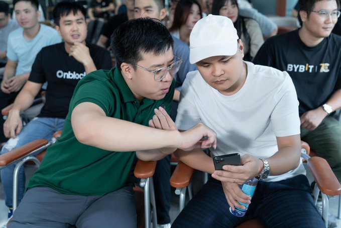 Two young men sit next to each other in an audience, looking at a smartphone; the man on the right wears a white cap, and the man on the left wears glasses and a green polo shirt.
