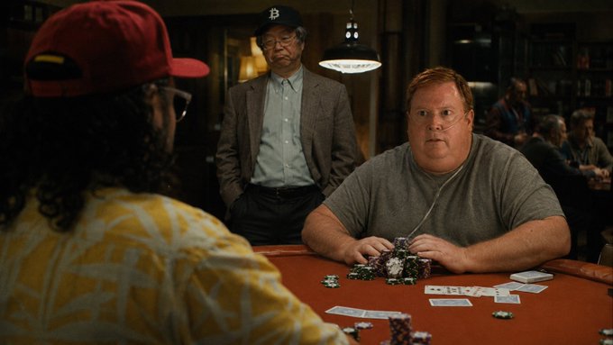 A scene from a film showing three men around a poker table, with a large man in a gray t-shirt facing two others, one wearing a red cap and another older man in a gray jacket and glasses.