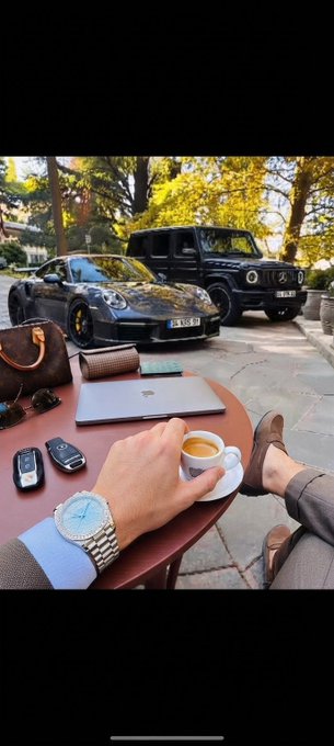 A first-person perspective showing a man holding an espresso cup while wearing a luxury watch, with a Porsche, Mercedes G-Wagon, designer bags, and a laptop arranged on a table.