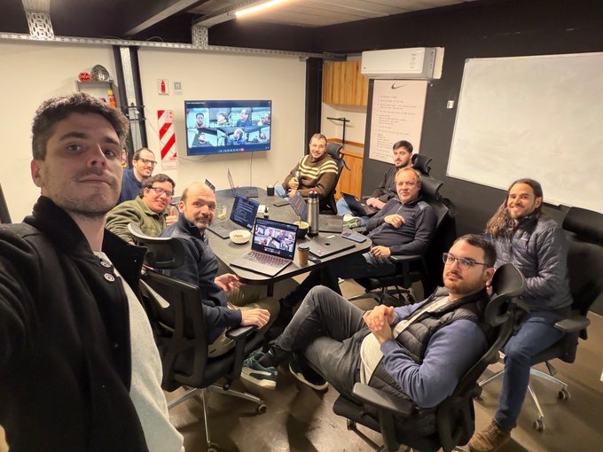 A group of nine developers poses for a selfie in a casual meeting room, smiling around a conference table with laptops and a wall-mounted screen displaying a video call, illustrating the team's collaborative effort behind the @ethrex_client project with @class_lambda, which was shared by @VitalikButerin.