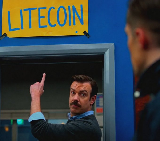 A man with a mustache points upward at a yellow "LITECOIN" sign taped to a blue wall, while another person stands in the foreground. The scene appears to be from a TV show or film, with the man wearing a dark sweater over a light blue collared shirt.