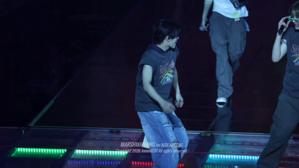 The image shows a stage with two people performing, one of whom is wearing a shirt that says "Shanahan" on it. The other person has their back turned towards the camera. There are also some lights visible in the scene, which may be part of the performance setup or lighting equipment for an event.