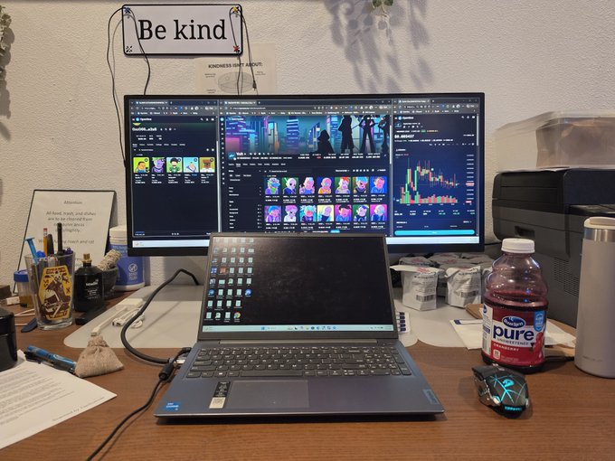 A home office desk setup with a laptop displaying trading charts and NFT collections on external monitors, a "Be kind" sign, a "pure" cranberry juice bottle, and a "Do your best" encouraging note.