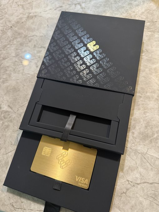 Photograph of a black card packaging box with metallic 'iBLOCK' patterned designs, partially opened to reveal a gold Visa-branded metal card being pulled out on a ribbon against a marble surface.