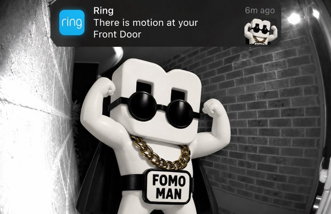 A 3D-rendered character wearing sunglasses and a 'FOMO MAN' chain necklace flexes its muscles in a black-and-white Ring doorbell camera feed, with a notification stating 'Ring: There is motion at your Front Door' timestamped '6m ago'.