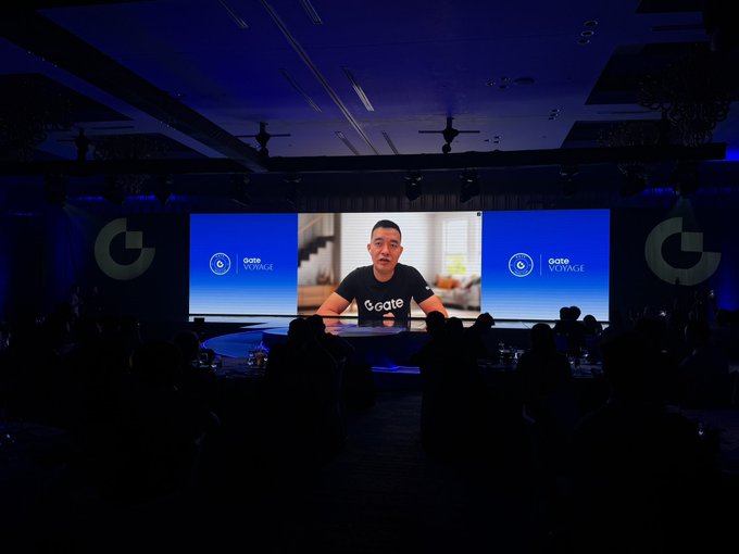 A view of a dark auditorium where a large screen displays a man speaking virtually, with the 'Gate VOYAGE' logo on both sides of the screen.