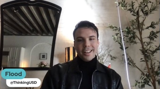 A screenshot shows a smiling man in a black leather jacket and turtleneck speaking on camera in a modern interior with a large mirror and leafy tree. A lower-third label reads “Flood” and “@ThinkingUSD,” matching the post’s “i came here for the comments.”