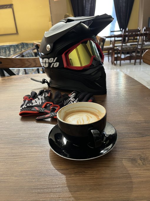 A black motorcycle helmet with yellow-tinted goggles marked "100%" sits beside a freshly poured cappuccino and riding gloves on a wooden table, suggesting the rider is taking a quick coffee break before or after a Sunday ride. The scene captures a moment of recharge, tying into the post's invitation to connect with others actively pursuing their passions on a Sunday.