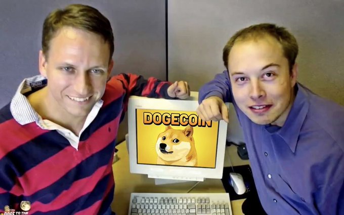 The image features two men standing next to a computer monitor, with one of them holding up a sign that displays "Dogecoin". The other man is smiling at the camera while they pose for the photo. The background includes a desk area with a keyboard and mouse on it.