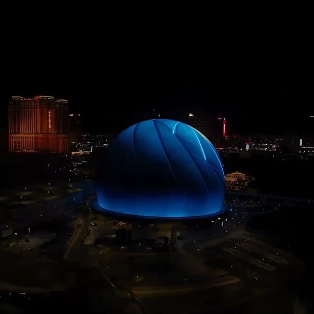 An aerial nighttime photograph of a large, illuminated blue spherical building in Las Vegas, with the glowing orange Encore hotel visible in the background.