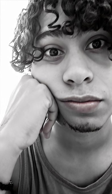 A close-up black-and-white portrait of a young man with curly hair, a goatee, and a ring on his finger, resting his chin on his hand.
