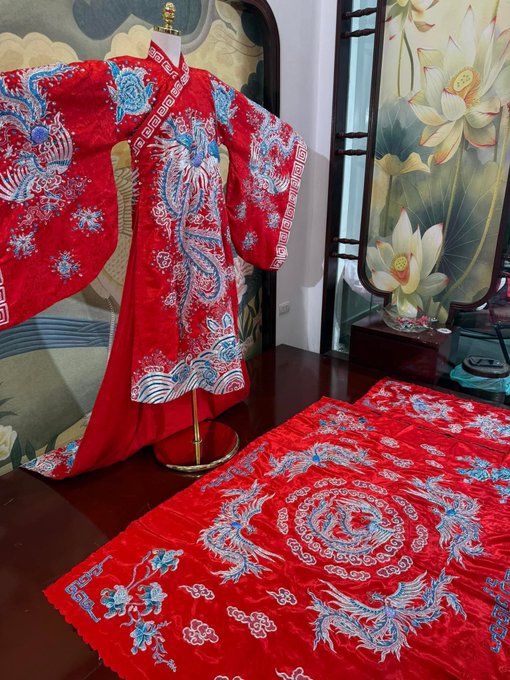 An ornate red traditional Chinese or Japanese-style robe with blue and white phoenix and dragon embroidery displayed on a mannequin, with matching embroidered fabric laid out on a wooden floor in front of a lotus flower painting.