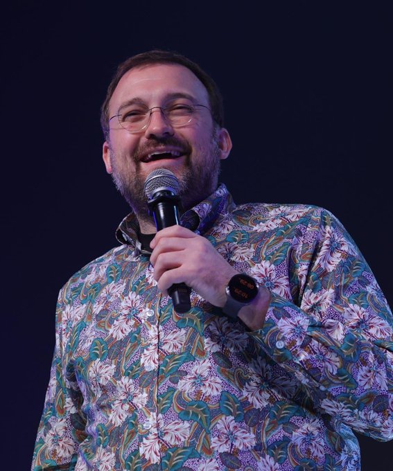 A photo of a man with glasses and a beard, smiling and holding a microphone, wearing a colorful paisley-patterned shirt.