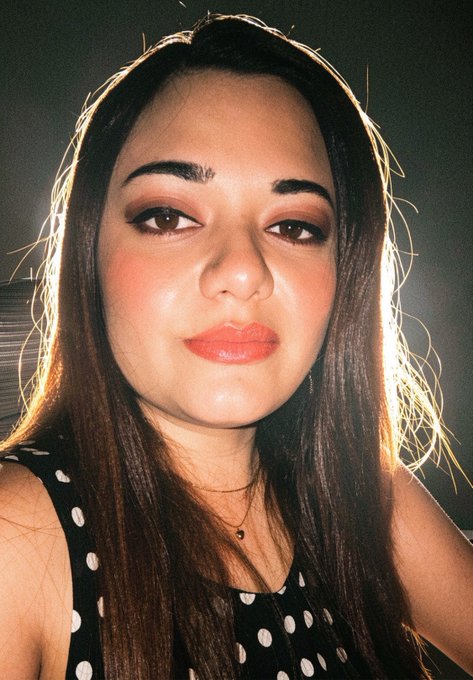 A close-up selfie of a young woman with long dark hair, winged eyeliner, and coral lipstick, wearing a black and white polka dot top and a delicate necklace.