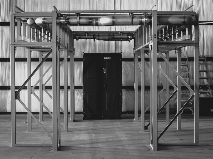 A black-and-white photograph of an industrial structure with metal scaffolding framing a large black electrical cabinet and overhead piping.