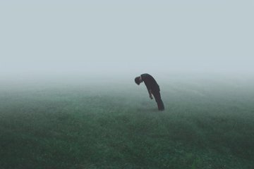 A solitary figure in dark clothing stands bent over in a misty, fog-covered field of green grass, conveying a sense of isolation and melancholy.
