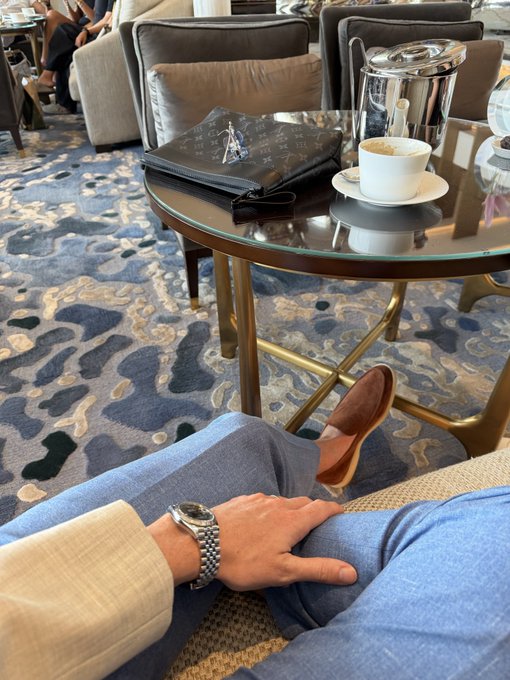 First-person view of a person in light blue trousers and brown loafers relaxing in a lounge, with a wristwatch visible, a Louis Vuitton pouch, coffee cup, and glass kettle on a table ahead.
