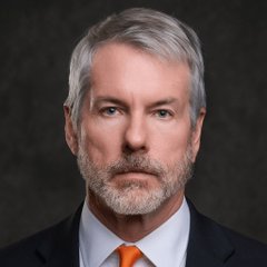 A portrait of a man with gray hair and a beard, wearing a dark suit and orange tie, photographed against a dark background.