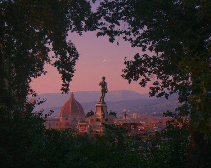 A crescent moon hangs over a statue silhouetted against a pink sunset sky, with the iconic dome of the Duomo di Firenze visible in the background, framed by dark tree branches—evoking the quiet anticipation of Florence at dusk, as if something is "soon" to come.