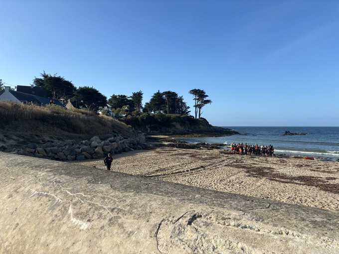 Beach scene with a group of people in orange jackets gathered near the shoreline, while one person in dark clothing stands alone on the sandy beach under a clear blue sky.