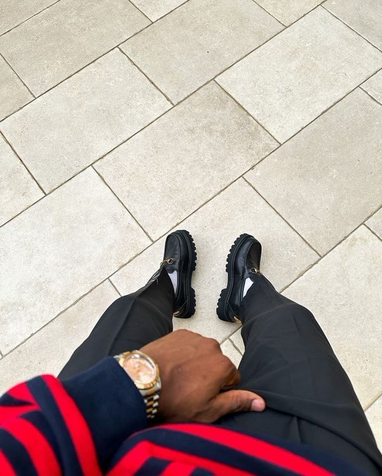 A first-person perspective photo looking down at a person wearing black pants, chunky black shoes, and a gold watch, with their hand resting on their leg against a tiled floor background.
