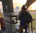 The image shows a man standing on top of a building, wearing safety gear such as a helmet and harness. He appears to be working at heights or possibly repairing something high up. The surrounding area seems to be an industrial setting with various equipment and structures visible in the background.