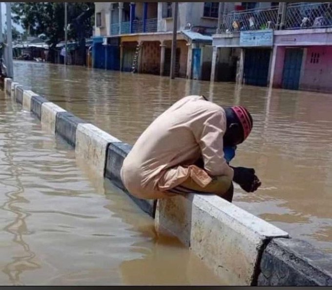 A person wearing a beige outfit and a red-and-black striped cap sits crouched on a flooded street divider, surrounded by murky floodwater that has inundated the surrounding buildings.