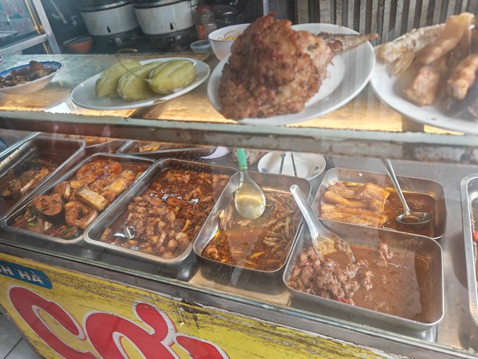 A display case shows various prepared dishes in metal trays, with fried foods and pickles on plates above, behind a yellow sign with red lettering.