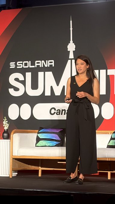 A woman in a black sleeveless jumpsuit speaking on stage at the 'SOLANA SUMMIT' with a 'Can...' visible on the backdrop, with a couch featuring colorful pillows behind her.