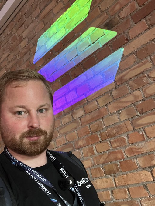 The image features a man wearing a black shirt with a lanyard around his neck. He has a beard and is standing against a brick wall. The text of the post reads, "GM What is the best wallet on @solana, And why is it @solflare?" This suggests that the man might be discussing or comparing different wallets available for purchase at the Bitcoin Cash exchange.