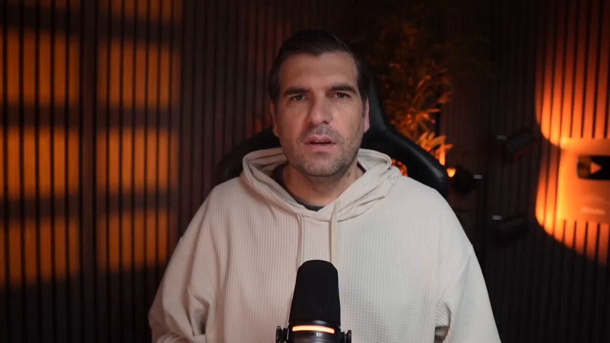 The image shows a man wearing a white hoodie holding a microphone with a "XRP" logo on his shirt. He appears to be speaking into the microphone, possibly discussing the current state of Ripple Coin or crypto currency. The background features a dark room with orange lights, which could represent an indoor setting like a recording studio or a dimly lit event space.