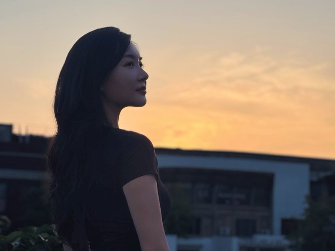 A young woman with long dark hair in a pensive pose, silhouetted against a vibrant orange sunset sky with buildings visible in the background.
