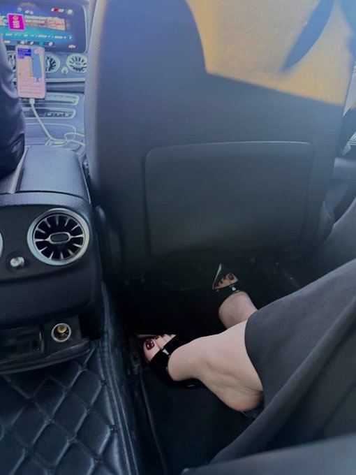 A person's feet in black sandals with red nail polish rest on the back seat floor of a car, near a charging phone.