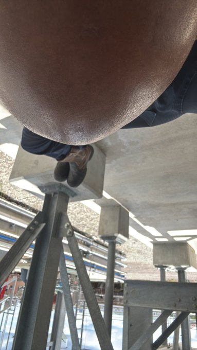 An upside-down selfie showing a person wearing a brown hat and jeans, standing on a metal staircase or scaffolding structure in an industrial setting.
