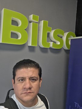 The image features a man wearing a backpack and standing next to a gray wall with a green "Bitsc" sign above him. The man appears to be looking at the camera, possibly posing for a photo or participating in an event related to cryptocurrency. The background of the image is blurred, drawing attention to the man and his surroundings.