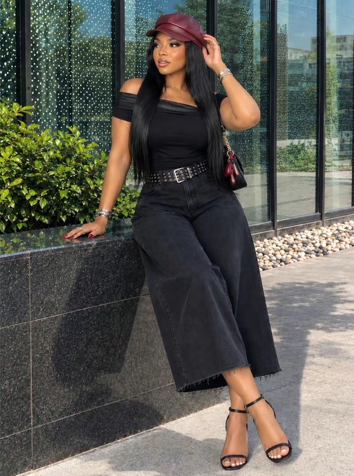 A woman with long dark hair poses outdoors wearing a burgundy cap, black off-shoulder top, studded belt, wide-leg black jeans, and strappy heels, carrying a quilted bag.