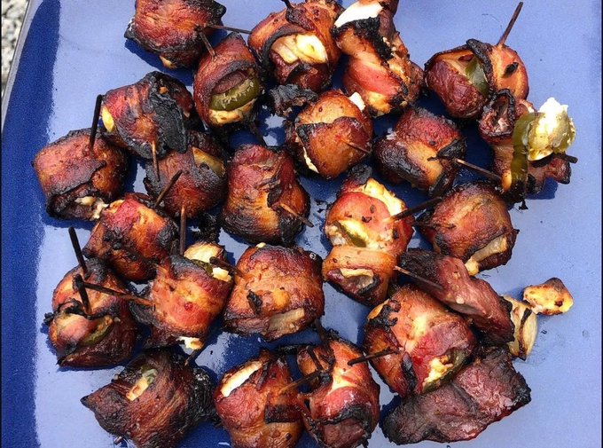 A close-up of bacon-wrapped appetizers, likely stuffed with jalapeños and cream cheese, secured with toothpicks and cooked on a blue tray.