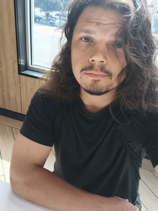 A man with long, wavy hair and a goatee sits wearing a black t-shirt, giving a calm, direct gaze to the camera in what appears to be a modern café or restaurant near a window. His serious, no-nonsense expression aligns perfectly with the post's message of working quietly and preferring no small talk.