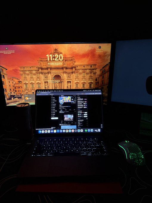 A MacBook displaying messaging and scheduling apps sits open in front of a large monitor showing a sunset skyline (time: 11:20, music: Angel's), accompanied by a glowing honeycomb-style mouse and a cylindrical speaker. The setup captures the "laptop, some SOL, & a dream" vibe—a sleek, after-hours workspace ready for trading or building into the night.