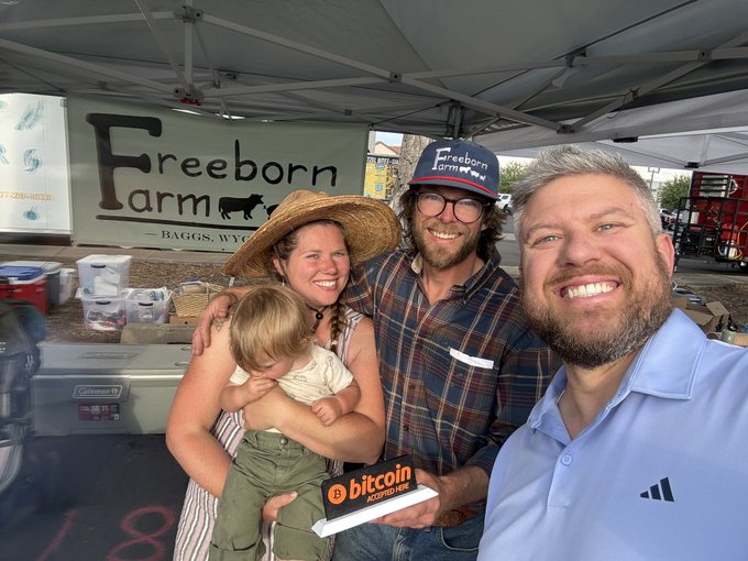 A selfie of three adults and a child at a farmer's market stand for Freeborn Farm (Baggs, WY), with one person holding a Bitcoin Shrimp Hat book.