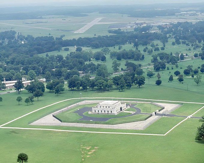 Aerial view of a large building with an octagonal wall surrounding it, set in a green field with trees and what appears to be an airstrip in the distance.