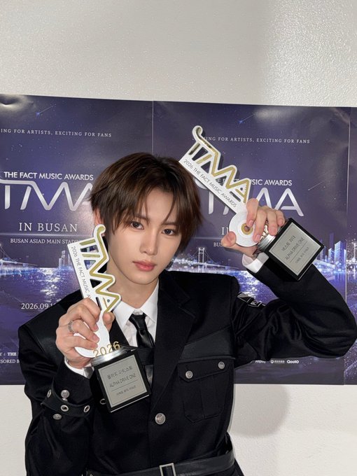 A young man with brown hair, dressed in a black suit and tie, poses with two trophies at "2026 THE FACT MUSIC AWARDS" (TMA) held in Busan. The hashtags reference Alpha Drive One (AL1), and the image appears to celebrate his win at the awards ceremony.