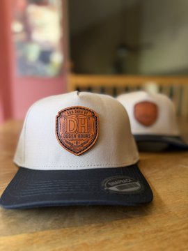 The image features two hats, one white with a brown leather patch that says "Dh" on it, and another black hat placed next to it. The hats are displayed on a wooden table or countertop, possibly at a store or an event related to the "Dh Days of Degen Hours". The hats appear to be handmade, as indicated by the text in the image.