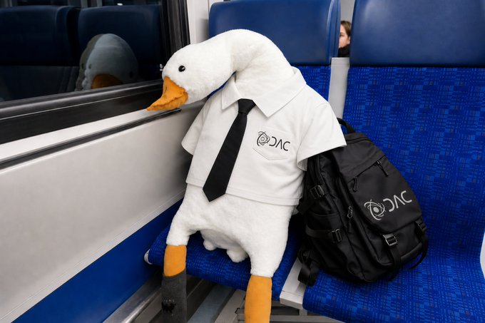 A white duck plush wearing a shirt and black tie, both branded with the "DAC" logo, sits on a blue train seat next to a matching DAC backpack, apparently gazing out the window. The image cleverly personifies DAC as a watchful, suited traveler—tying into the post's portrayal of @dac_chain as a vigilant, prepared player in addressing the quantum threat, rather than treating it as a distant issue.