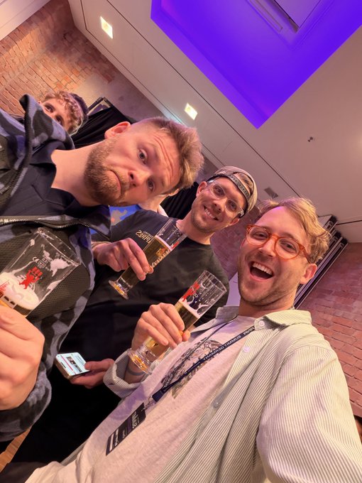 The image shows a group of four men standing together, each holding up a glass filled with beer. They are all smiling and posing for a photo, creating an atmosphere of camaraderie and enjoyment. The man at the center of the frame is holding his glass in front of him, while the other three men have their glasses raised as well. The background features a brick wall and a purple ceiling, adding to the overall ambiance of the scene.