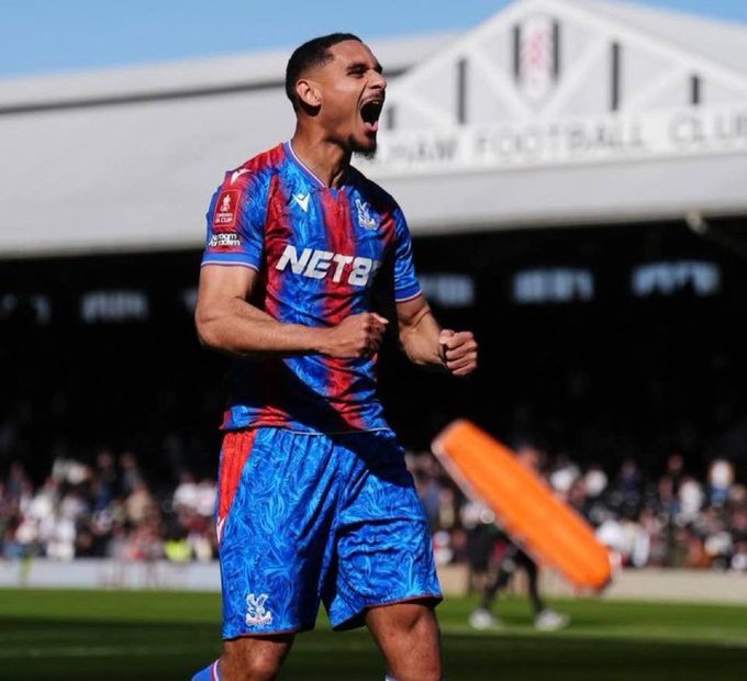 A soccer/football player in a blue and red Crystal Palace kit with 'NET8' sponsor celebrating with a raised fist, shown on a football pitch with stadium in the background.