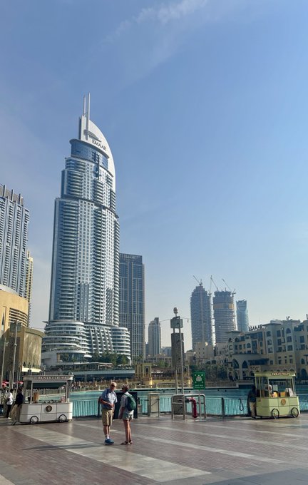A daytime photo of a tall modern skyscraper with twin spires in a city, with two people standing in the foreground near what appears to be a waterfront promenade.