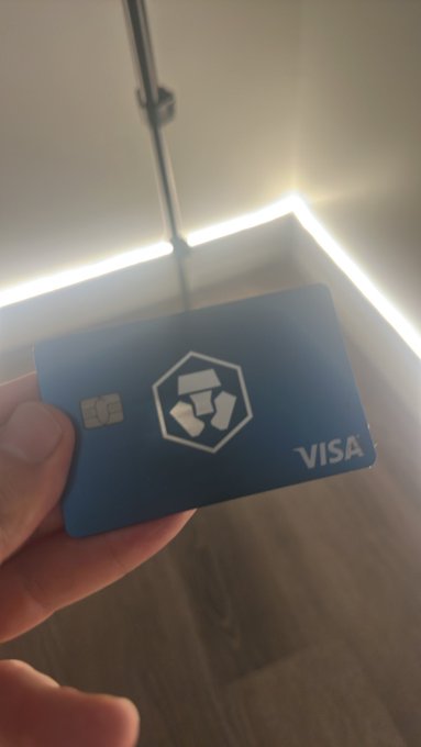 A hand holds a blue Crypto.com Visa-branded debit card in front of an LED-lit mirror and wooden floor.