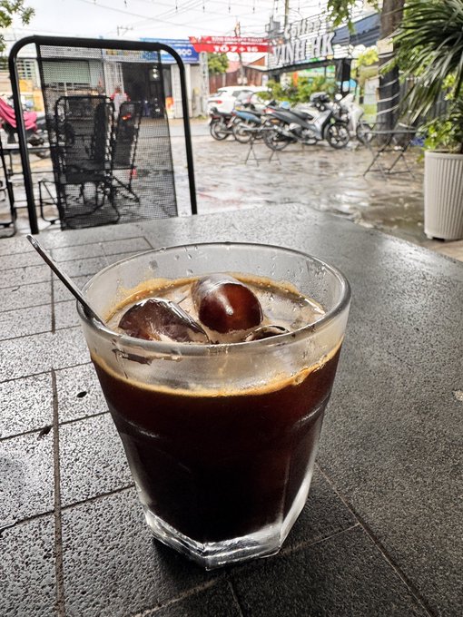 A close-up of a glass filled with iced coffee and spherical ice cubes sitting on a stone table, with a rainy street, parked motorcycles, and storefronts visible in the background.